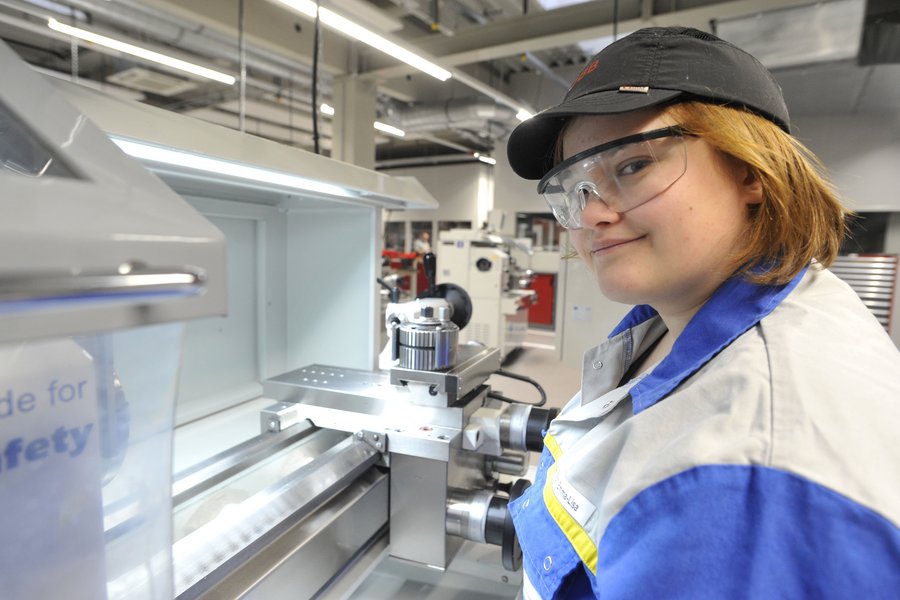 Eine junge Frau mit Käppi, Schutzbrille und Arbeitsanzug an einer Maschine