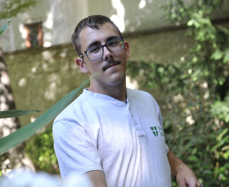 Ein junger Mann mit Schnauzbart und Brille steht inmitten von Grünpflanzen