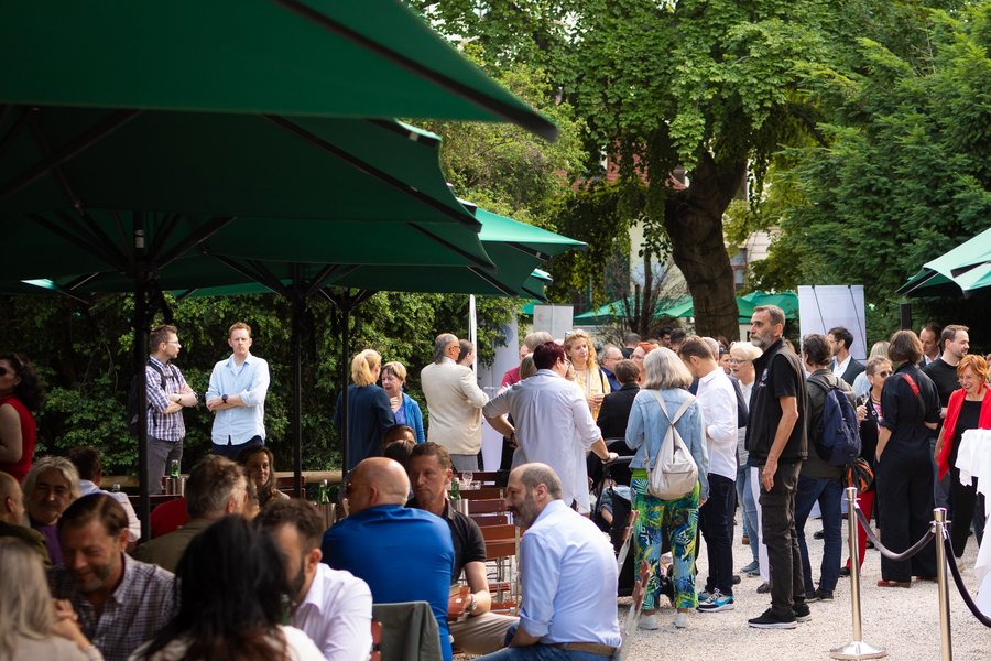 Viele Menschen essen, trinken und plaudern in einem Gastgarten mit großen grünen Sonnenschirmen.