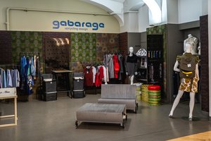 Ein gabarage-Geschäft: Im Hintergrund Kleiderstangen mit bunten Kleidungsstücken, im Vordergrund ein Sitzmöbel aus Rolltreppenstufen und eine Schaufensterpuppe mit gabarage-Rucksack