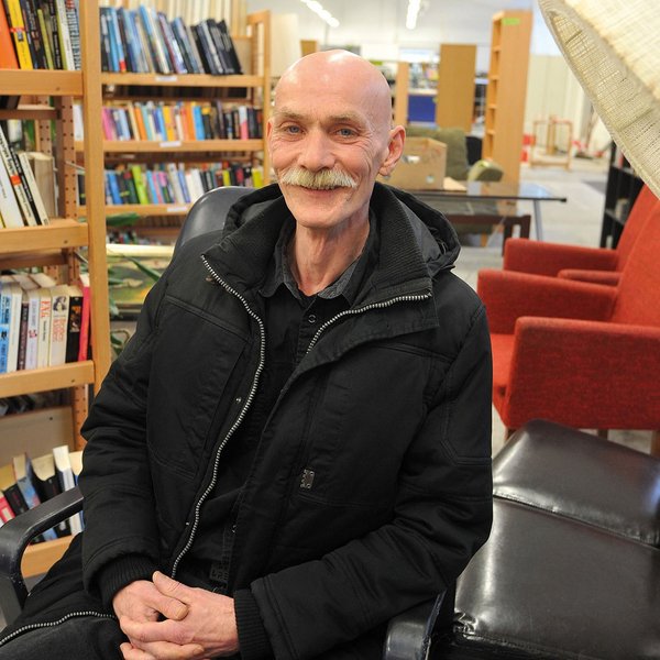Ein schlanker, Mann mit Glatze und Schnurrbart inmitten von Möbeln und Bücherregalen.