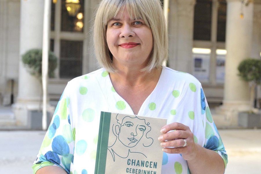 Eine Frau mit kinnlangen blonden Haaren und gepunktetem T-Shirt hält ein Buch in die Kamera