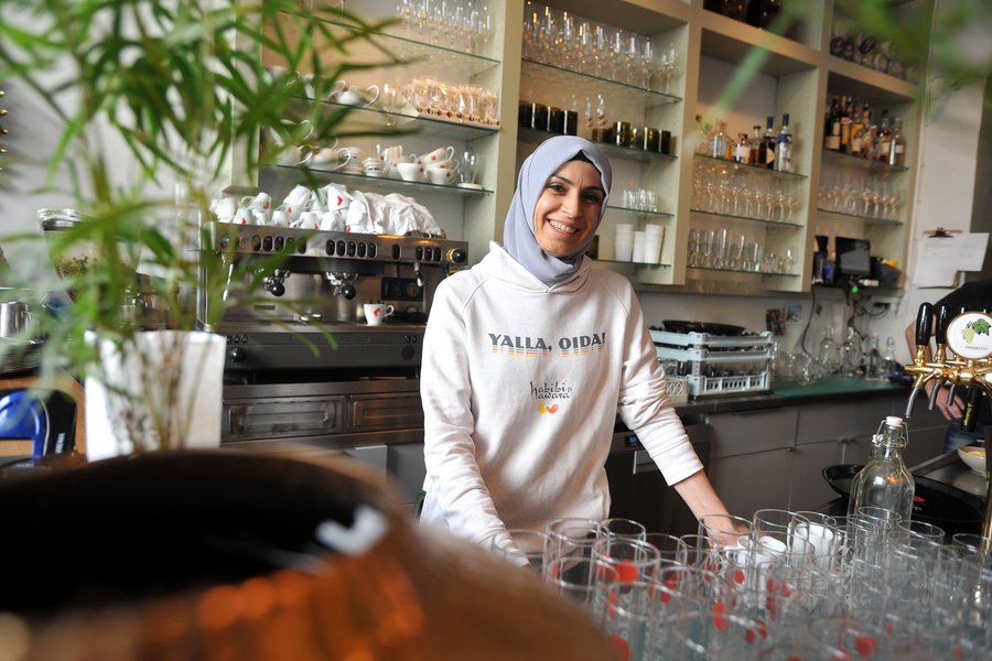Frau mit Kopftuch und Sweater steht lächelnd hinter einer Restaurantbar