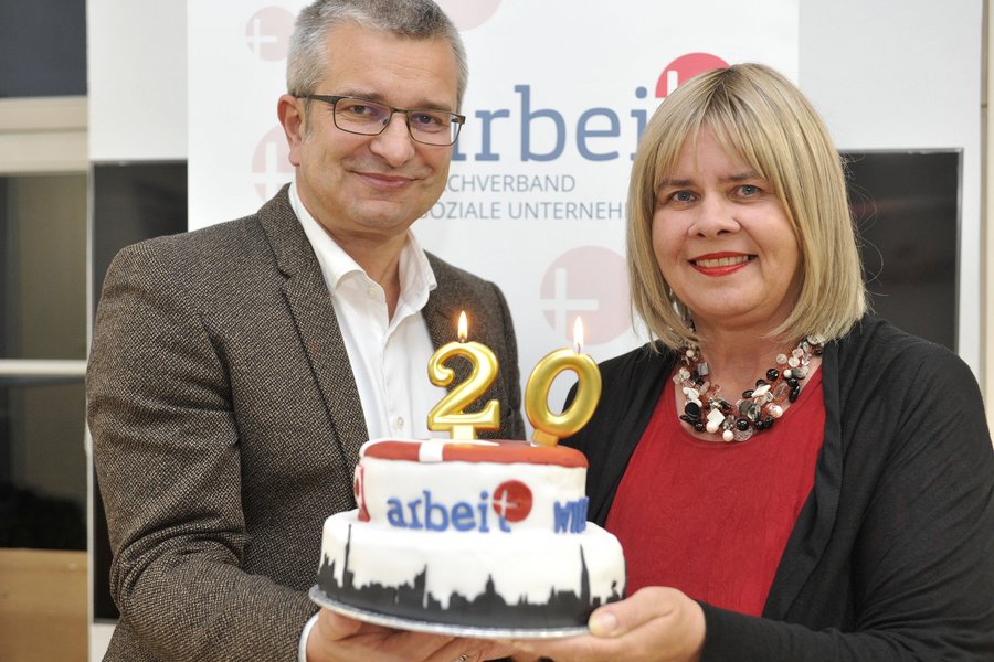 Ein Mann im Anzug mit Brille und sehr kurzen grauen Haaren und eine Frau mit schulterlangen Haaren präsentieren eine zweistöckige Torte mit Logo, Wien-Silhouette und 20er-Kerze